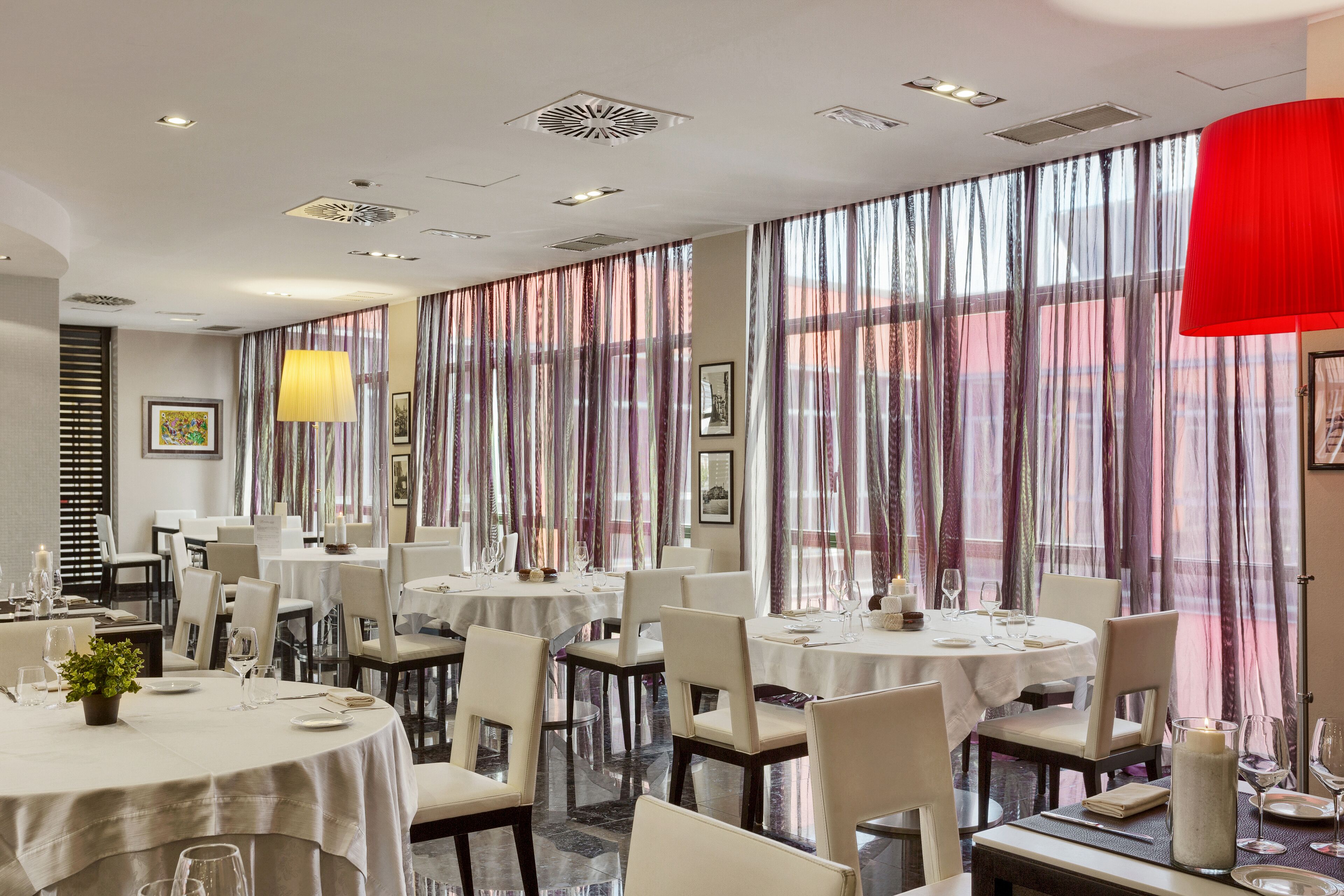 A restaurant with round tables, white chairs, and large windows with sheer curtains.