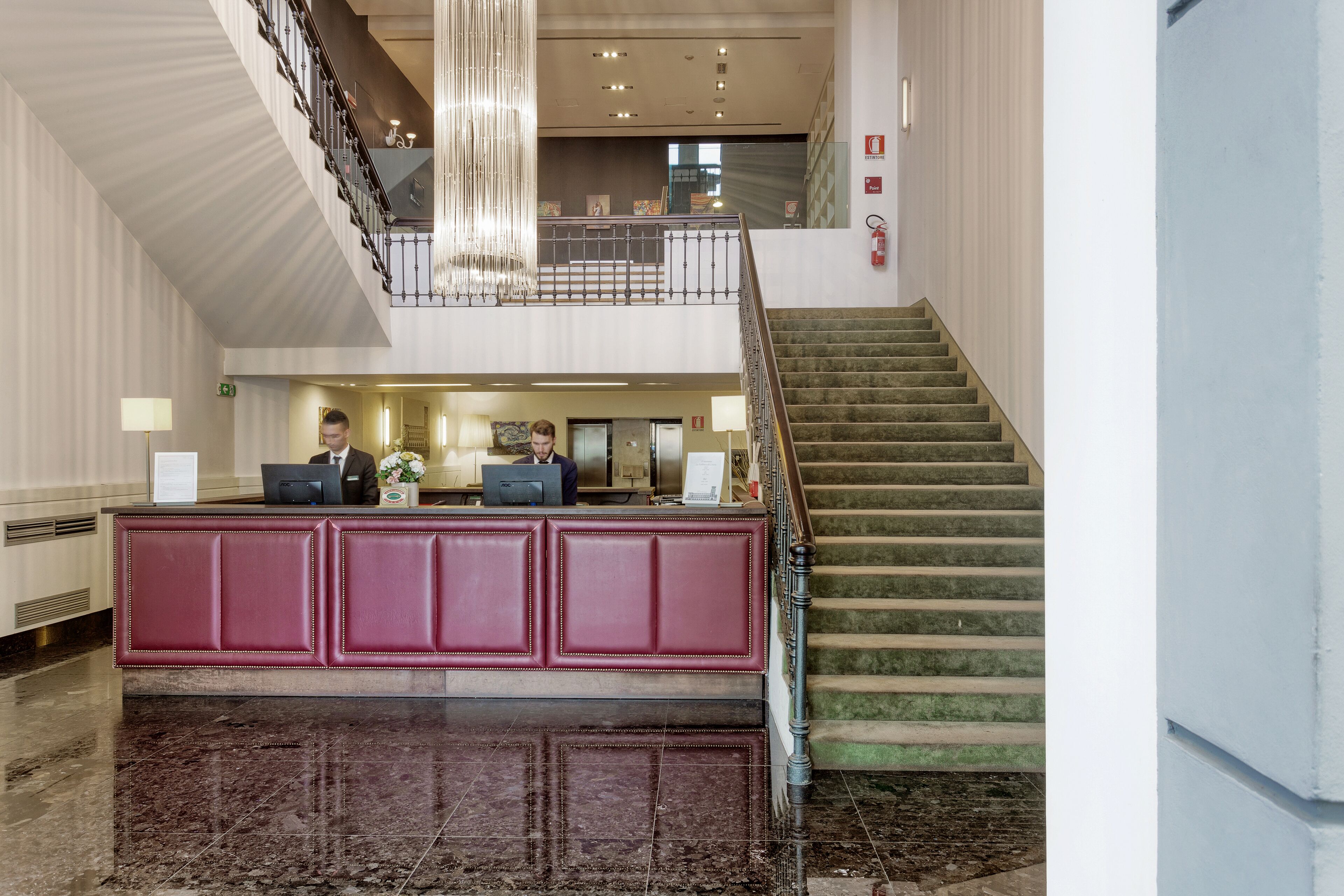 Een hotel receptie met een marmeren vloer, een rode receptiebalie en een trap naar een hoger niveau.