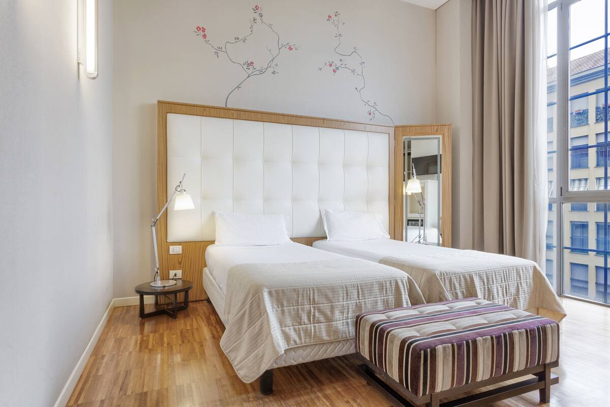 Une chambre d’hôtel avec deux lits, une table de chevet, une lampe, un miroir et une fenêtre donnant sur des immeubles.