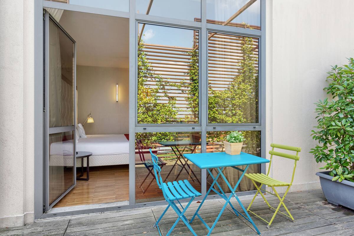 Un espace extérieur moderne aménagé avec une table bleue et des chaises vertes, une terrasse en bois et une vue sur un jardin, visible à travers de grandes fenêtres.