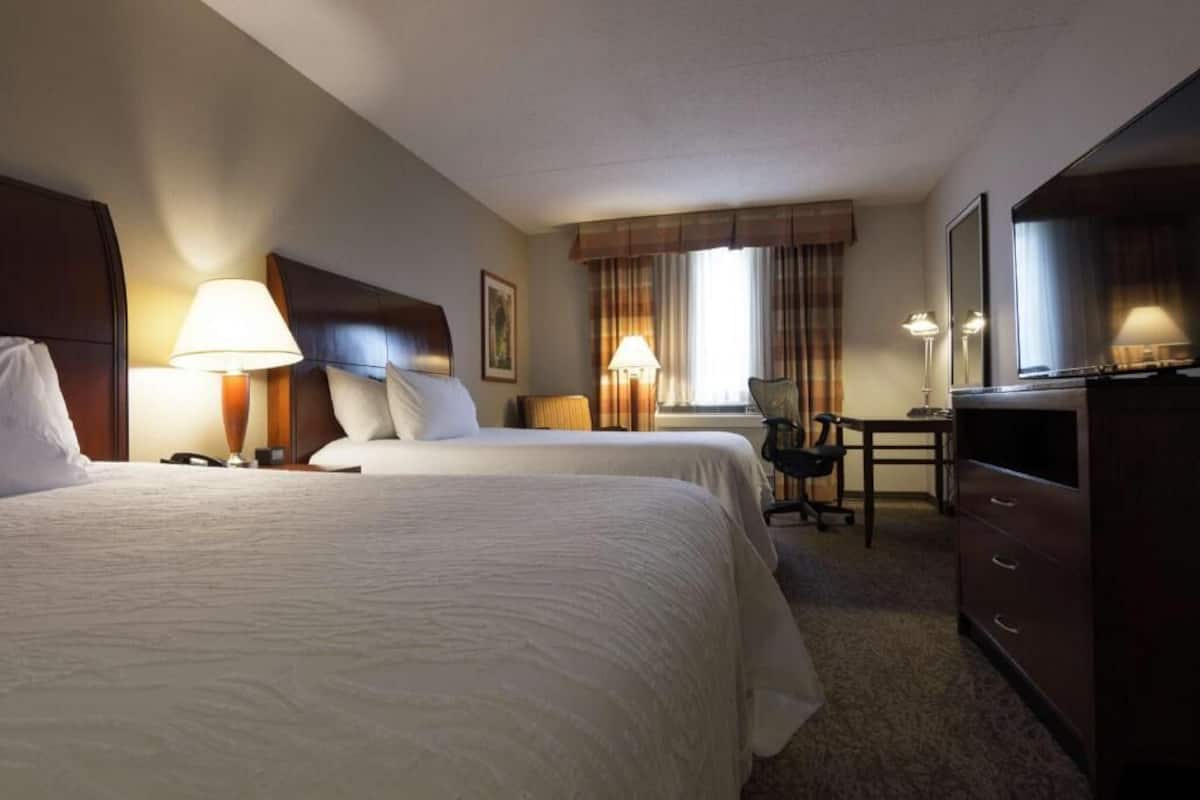 A hotel room with two beds, a desk, a chair, a television, and a lamp.
