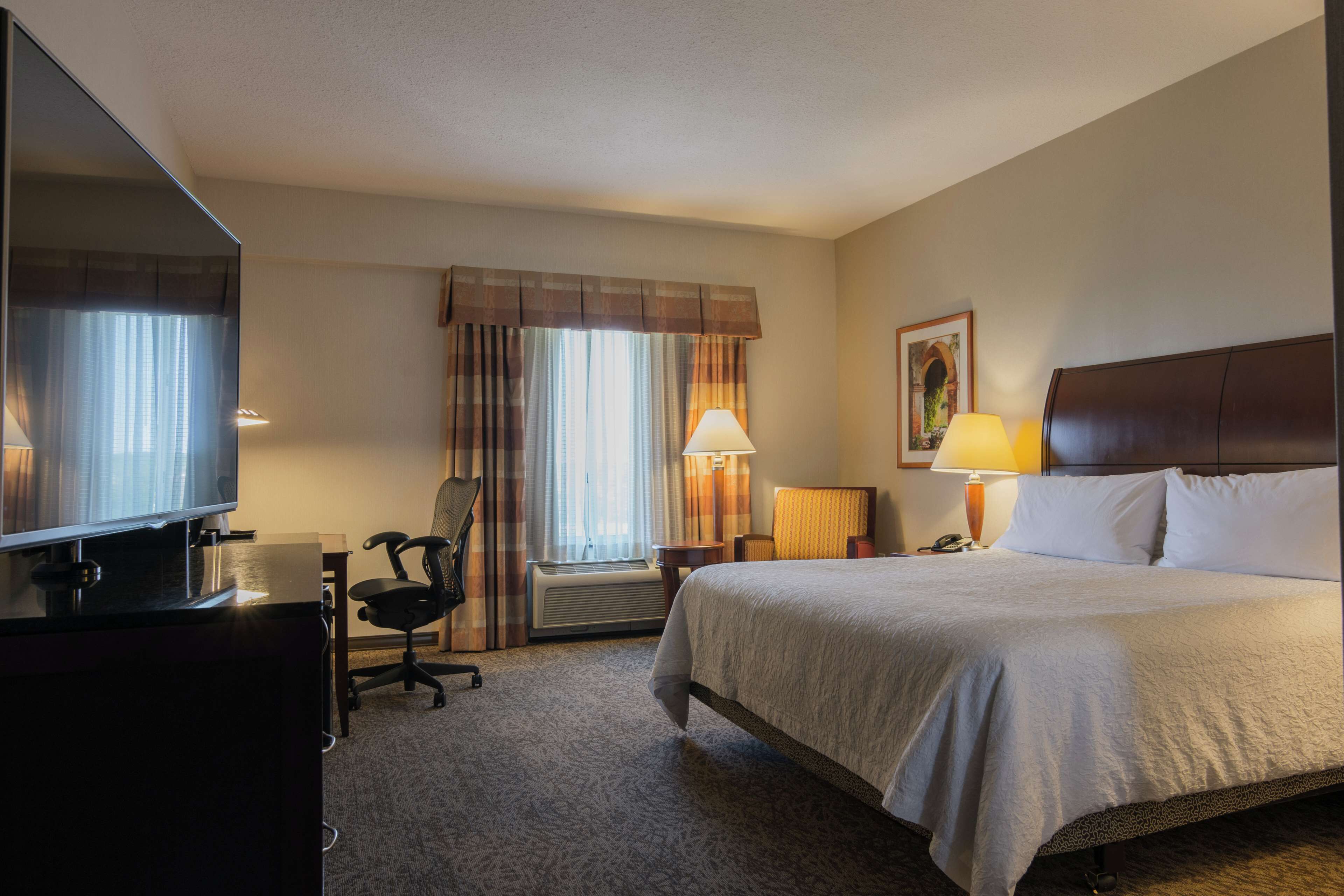 Quarto de hotel com cama, escrivaninha com computador, cadeira, televisão, abajur e janela com cortinas.