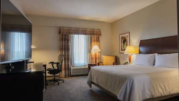 Quarto de hotel com cama, escrivaninha com computador, cadeira, televisão, abajur e janela com cortinas.