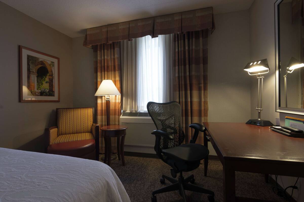 A hotel room with a bed, a desk, two chairs, a lamp, a mirror, and a window with curtains.