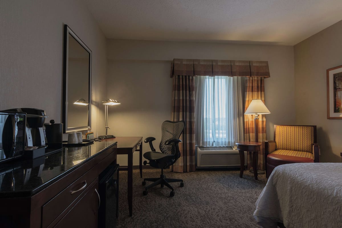 A hotel room with a bed, a desk, a chair, a mirror, a lamp, and a window with curtains.