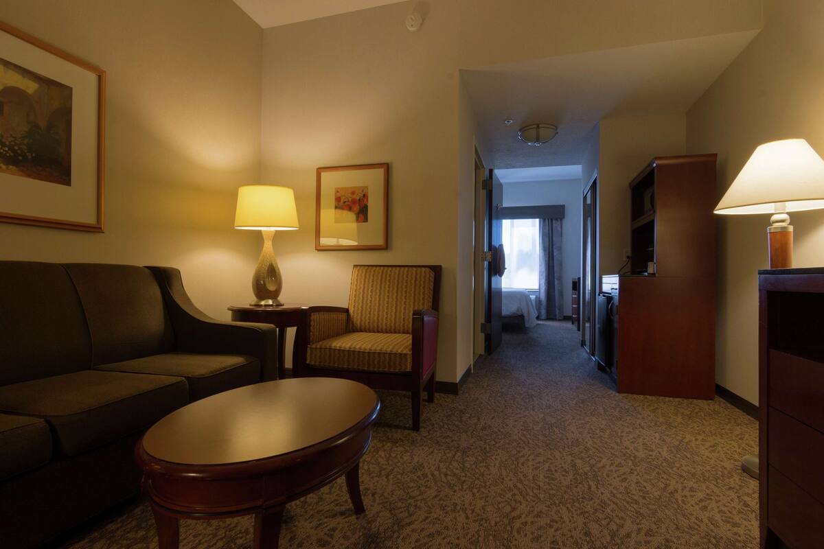 A hotel room with a sofa, armchair, table, and lamps. There is a hallway leading to a bathroom and a TV in the corner.