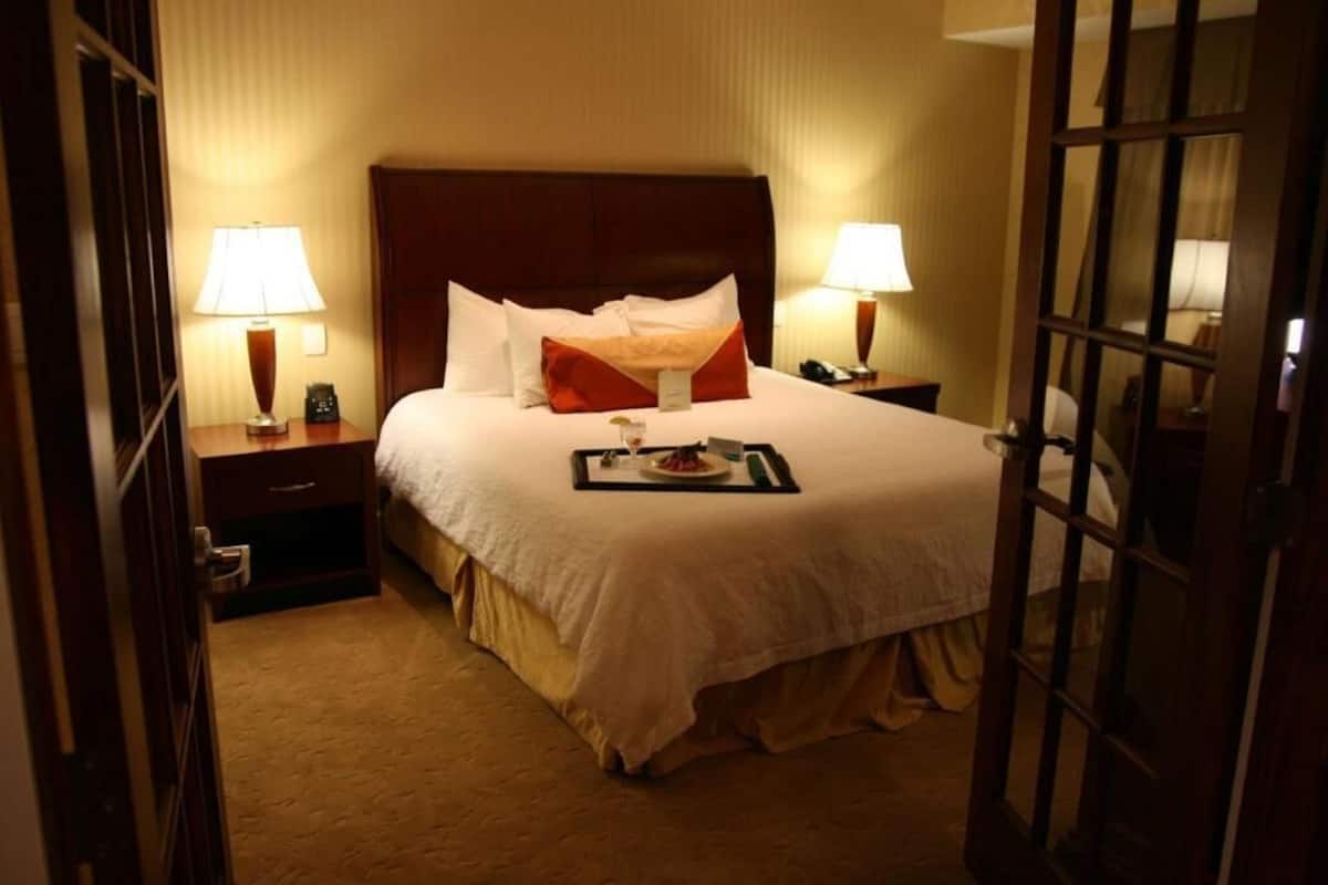 A hotel room with a large bed, two bedside tables with lamps, a nightstand, and a door.