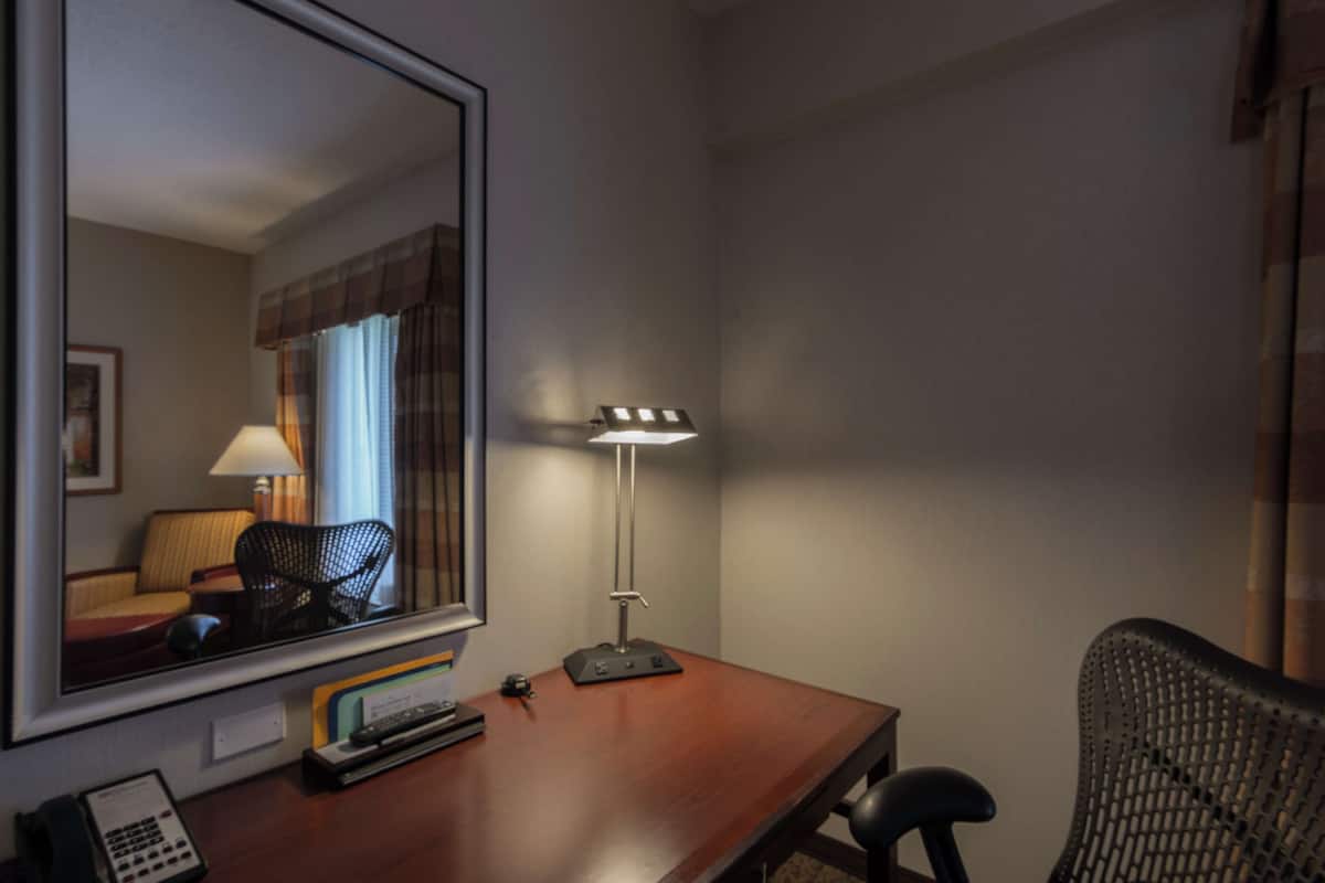 A hotel room with a wooden desk, a mirror, a lamp, a chair, and a telephone.