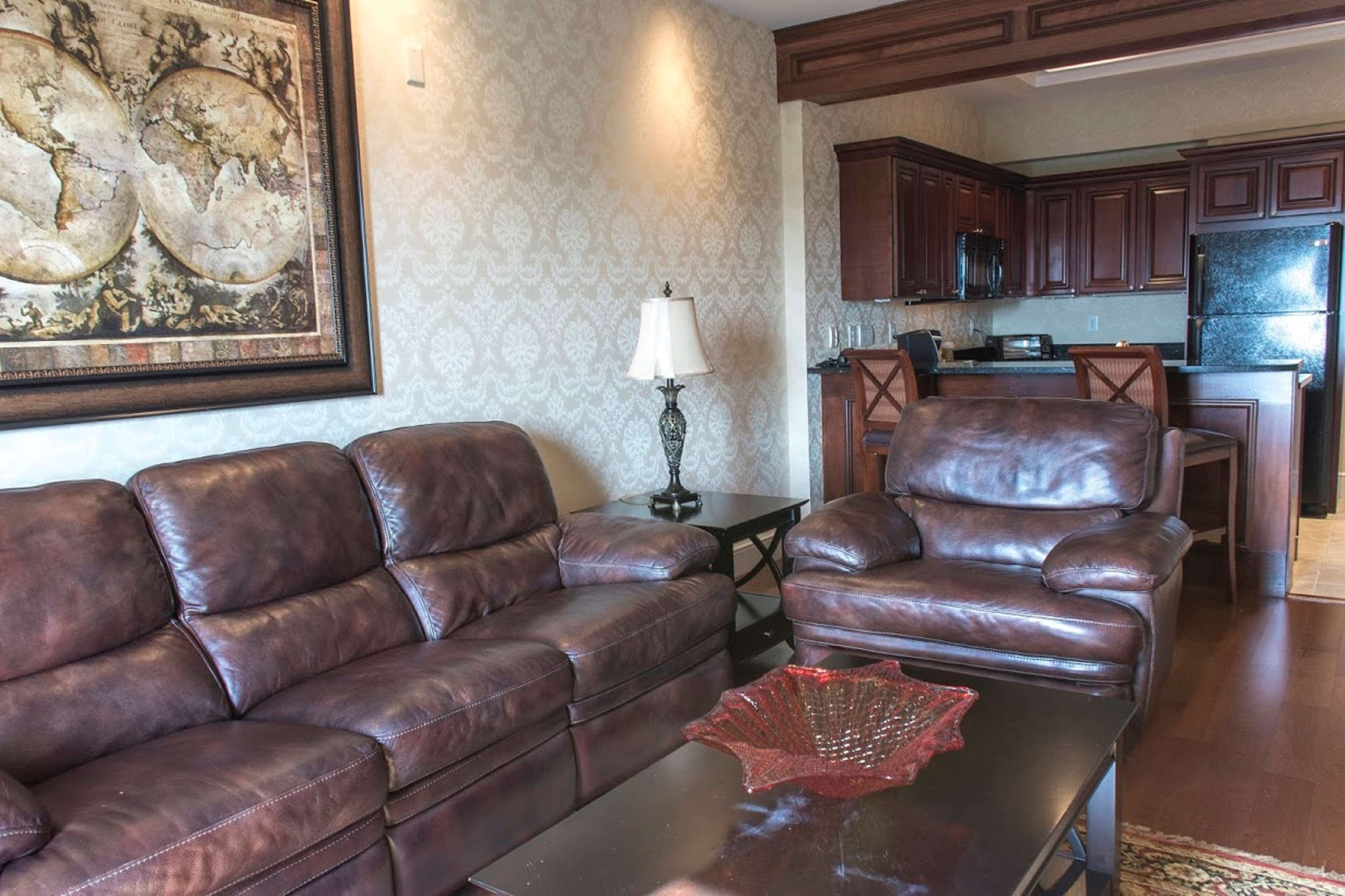 A living room with a brown leather sofa, a coffee table with a red decorative item, and a lamp. There is a kitchen area in the background with wooden cabinets and a refrigerator.