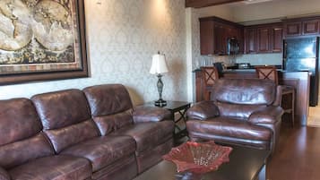 A living room with a brown leather sofa, a coffee table with a red decorative item, and a lamp. There is a kitchen area in the background with wooden cabinets and a refrigerator.
