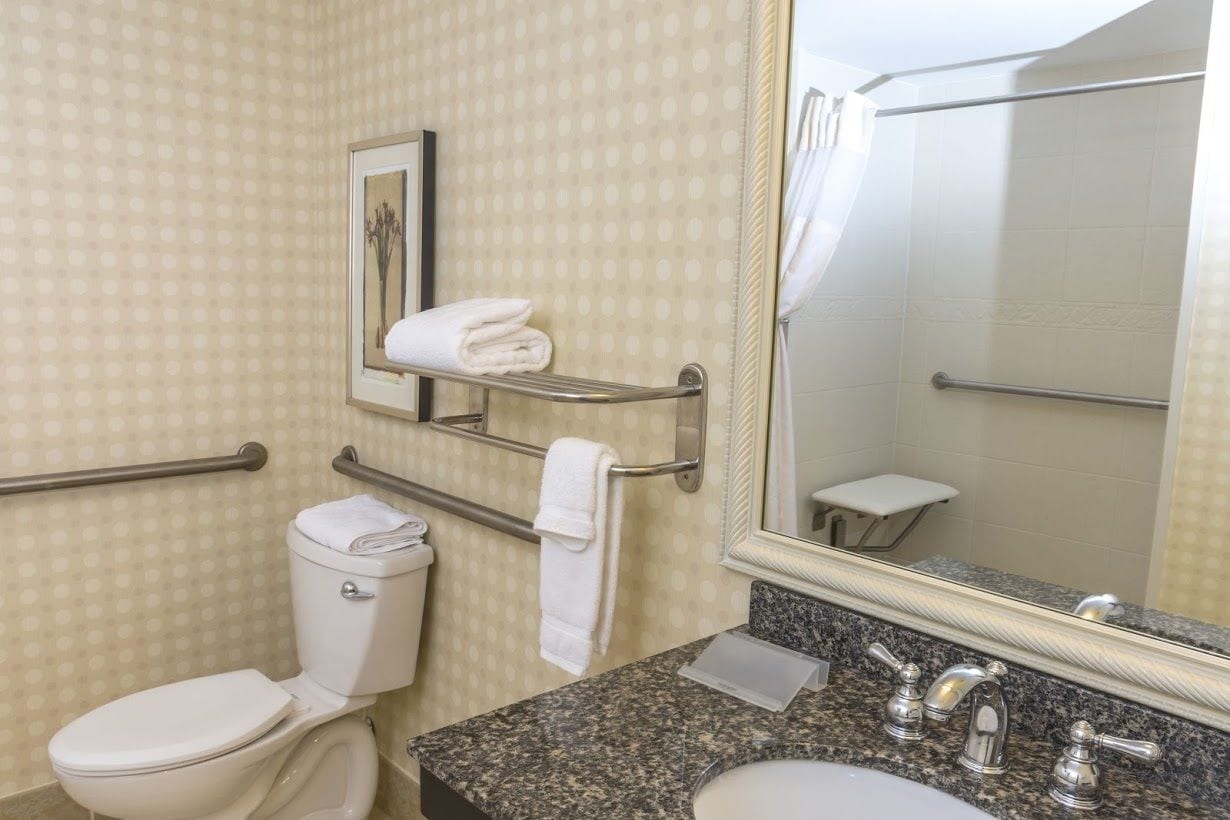 Une salle de bain équipée de toilettes, d’un lavabo et d’un miroir. On y trouve également des barres d’appui et un porte-serviettes.