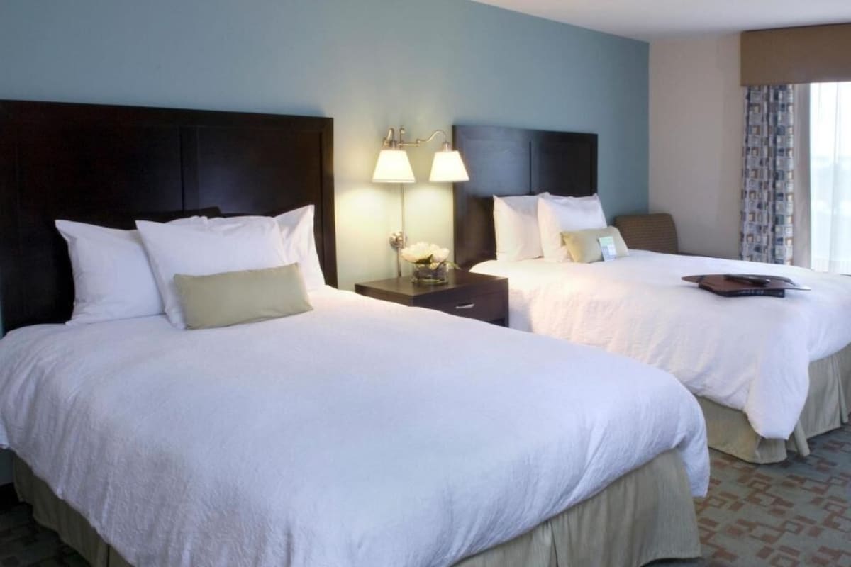 A hotel room with two beds, a nightstand with lamps, a chair, and a window with curtains.