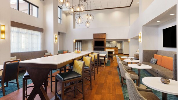 A modern hotel lobby with a long reception desk, multiple seating areas, and a kitchen in the background.