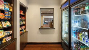A hotel pantry with shelves stocked with snacks and beverages, a refrigerator with various drinks, and a mirror reflecting the room.