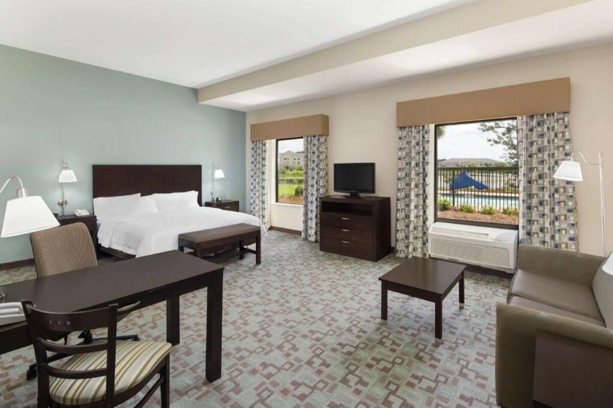A hotel room with a bed, a desk, a chair, a TV, a dresser, and a window with curtains.