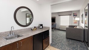 A compact hotel room with a kitchenette, a round mirror, a gray sofa, and a patterned carpet.