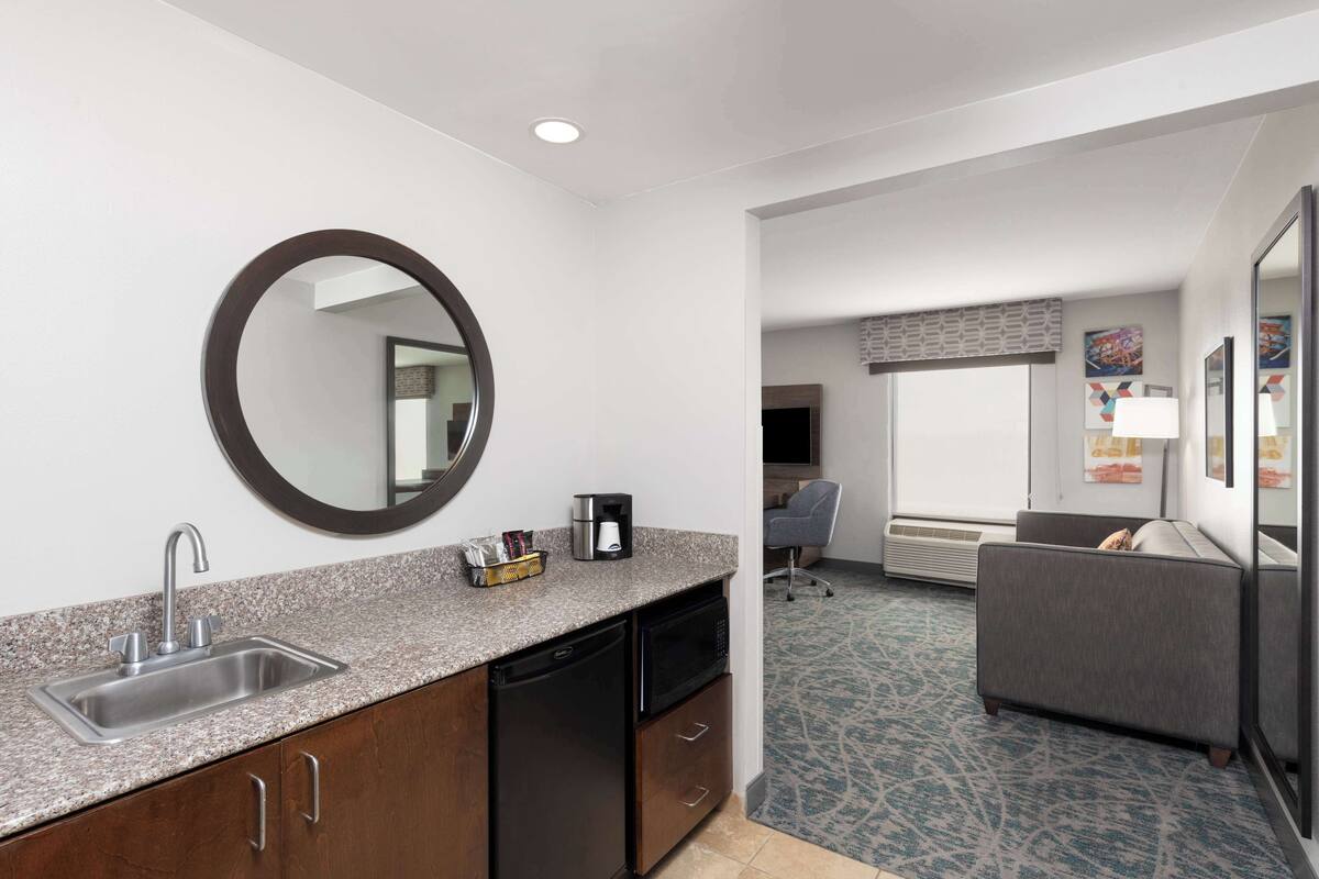 A compact hotel room with a kitchenette, a round mirror, a gray sofa, and a patterned carpet.