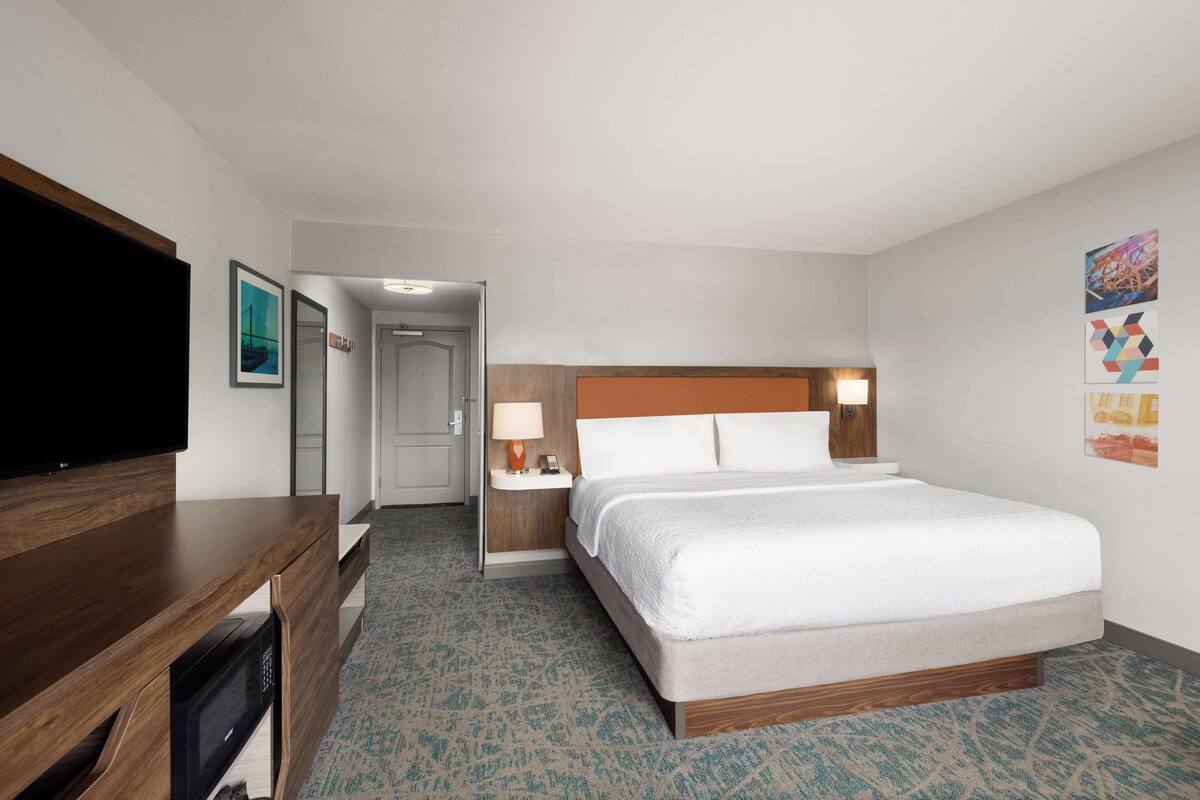 A hotel room with a bed, a desk with a flat-screen TV, a nightstand with a lamp, and a door leading to a hallway.