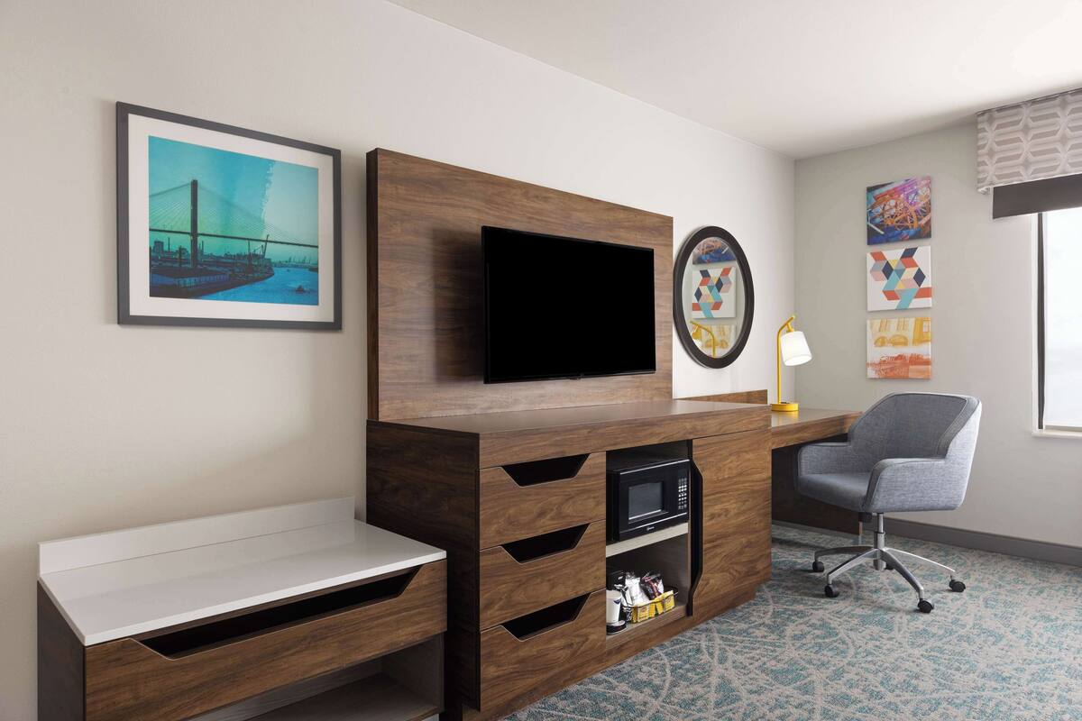 A hotel room with a wooden TV stand, a flat-screen TV, a desk, a chair, and a patterned carpet.