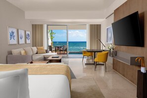 A hotel room with a bed, a sofa, a dining table with chairs, a TV, and a view of the ocean.