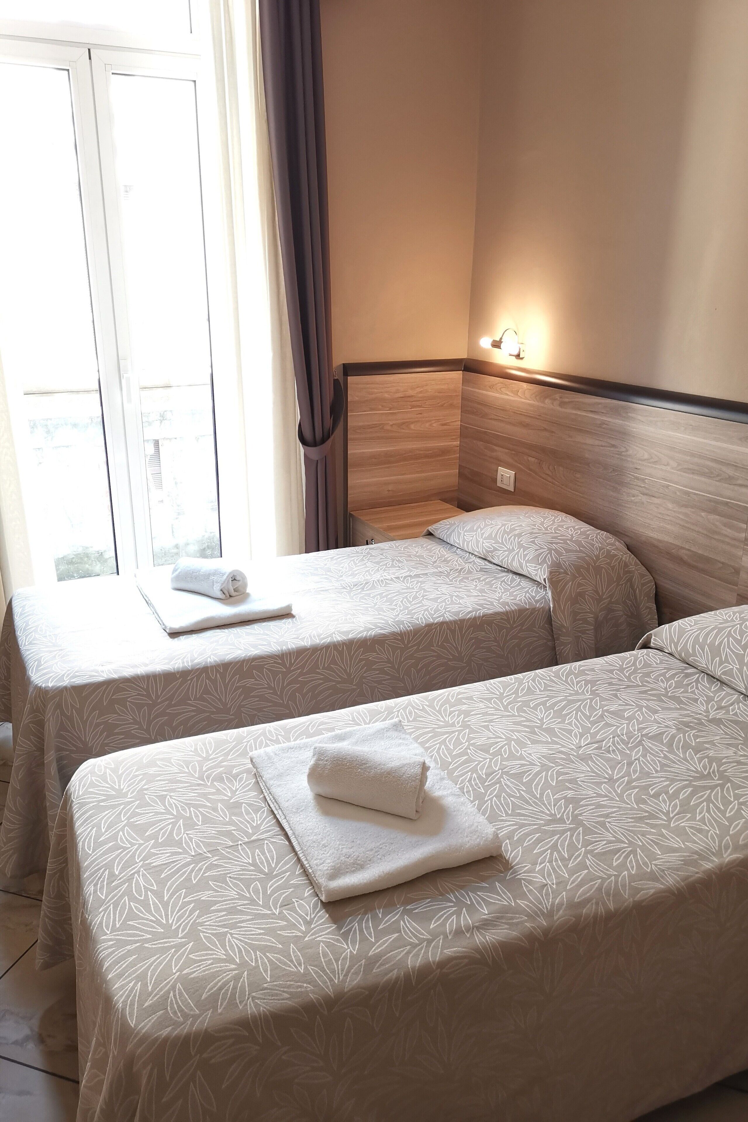 A hotel room with two beds, each with a white bedspread and a rolled towel on top. There is a window with curtains, a nightstand with a lamp, and a wooden headboard.