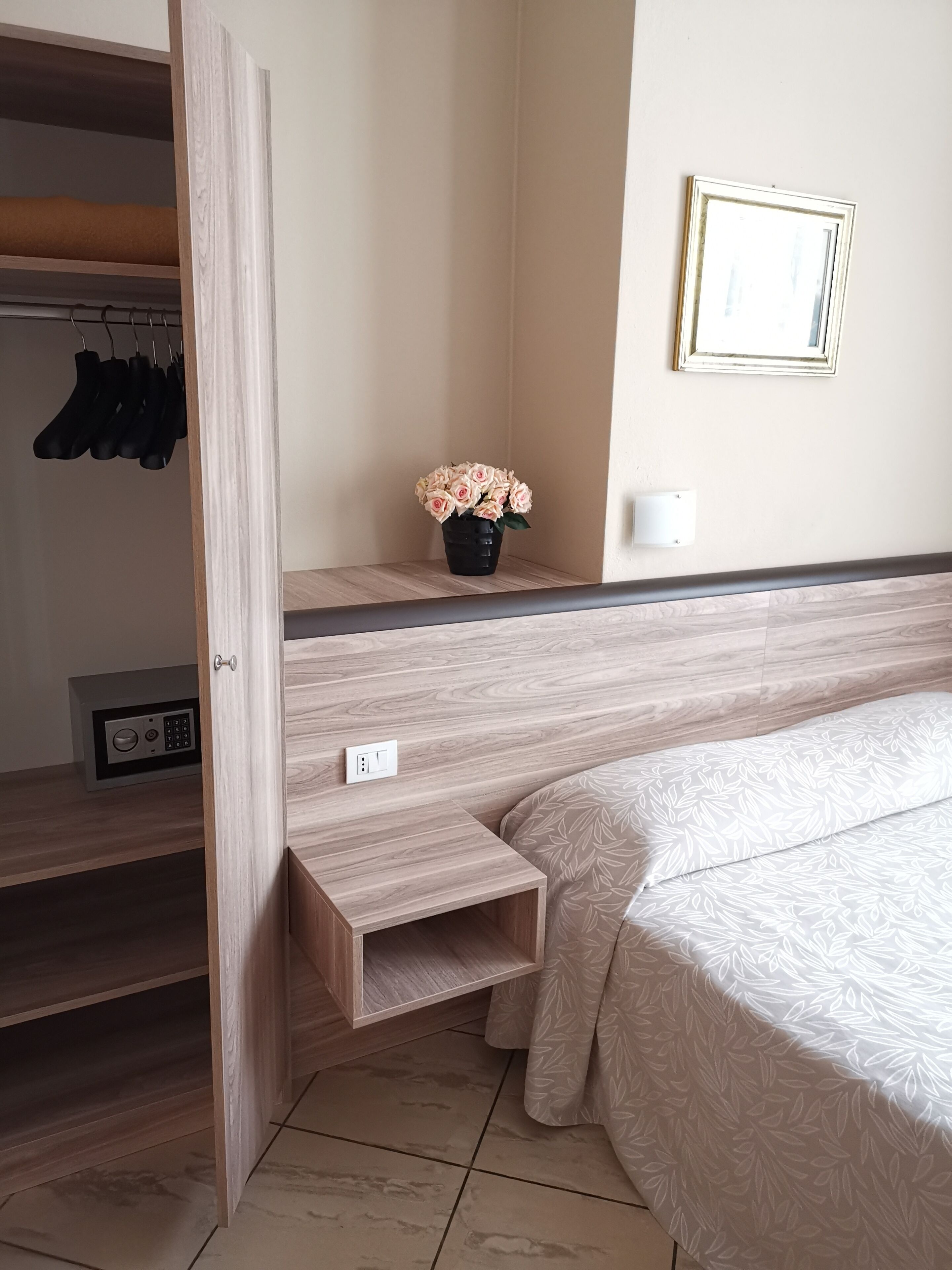A hotel room with a bed, a small bedside table, a wall-mounted light, a security keypad, a coat rack, and a potted plant.