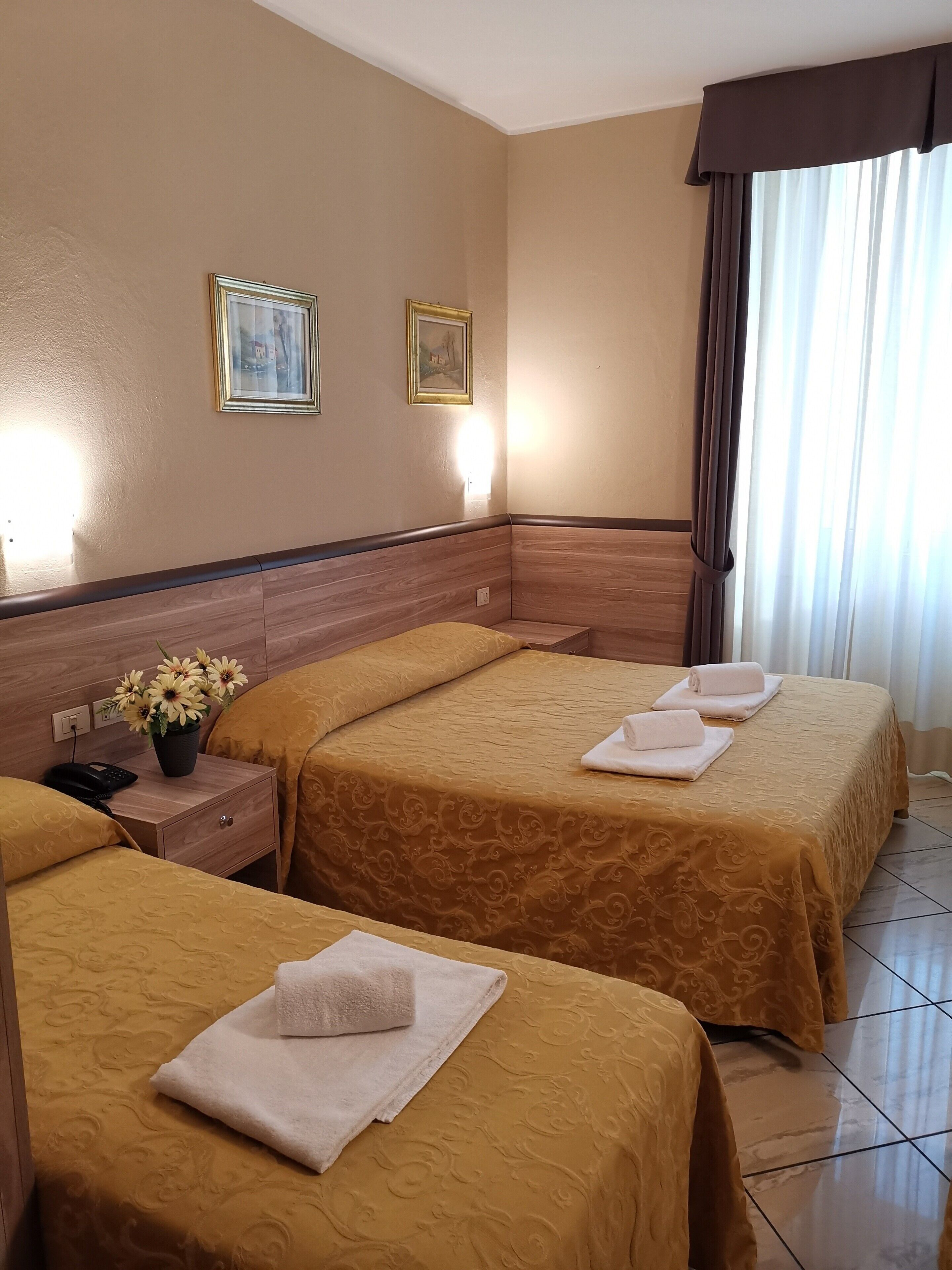A hotel room with two beds, a nightstand with a phone and a vase of flowers, and two framed pictures on the wall.