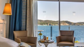 Habitación de hotel con dos sillas, una cama, una mesa con cosas para el desayuno y vistas al mar y a las colinas.