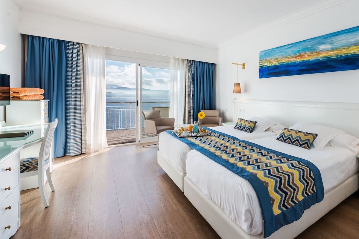 A hotel room with a large bed, a desk, a chair, and a view of the sea.