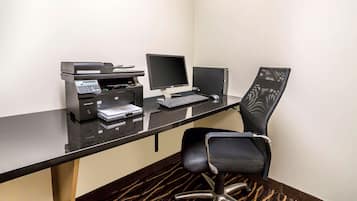 Escritório de hotel com uma mesa preta, uma impressora, um monitor, um teclado e uma cadeira.