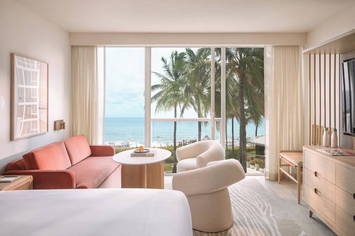 A hotel room with a large bed, a sofa, a chair, a small table, and a TV. The room has large windows with a view of the ocean and palm trees.