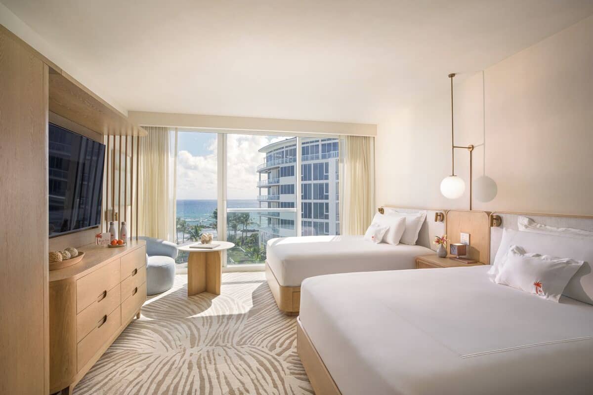 A hotel room with two beds, a TV, a minibar, a view of the ocean, and a large window.