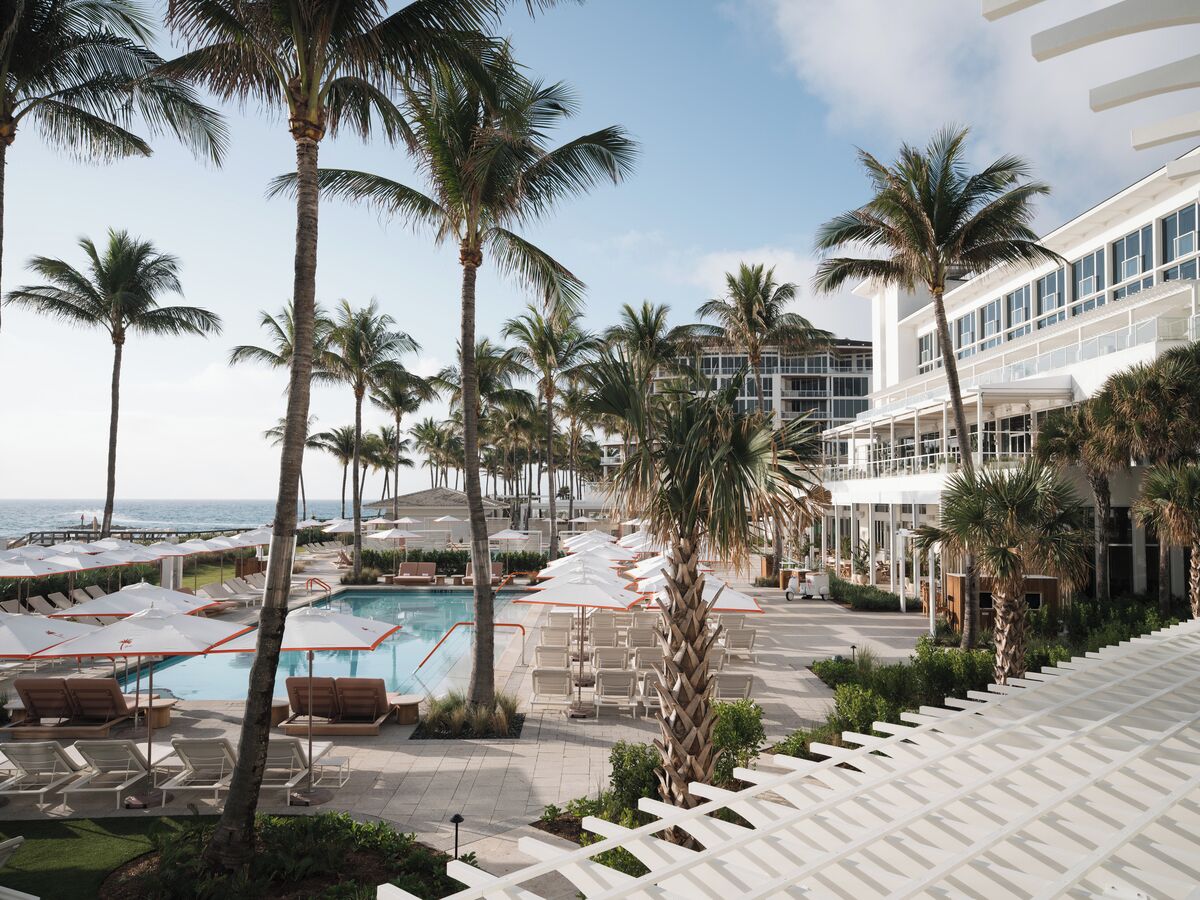 A resort pool area with sun loungers and umbrellas, surrounded by palm trees and a building with large windows.