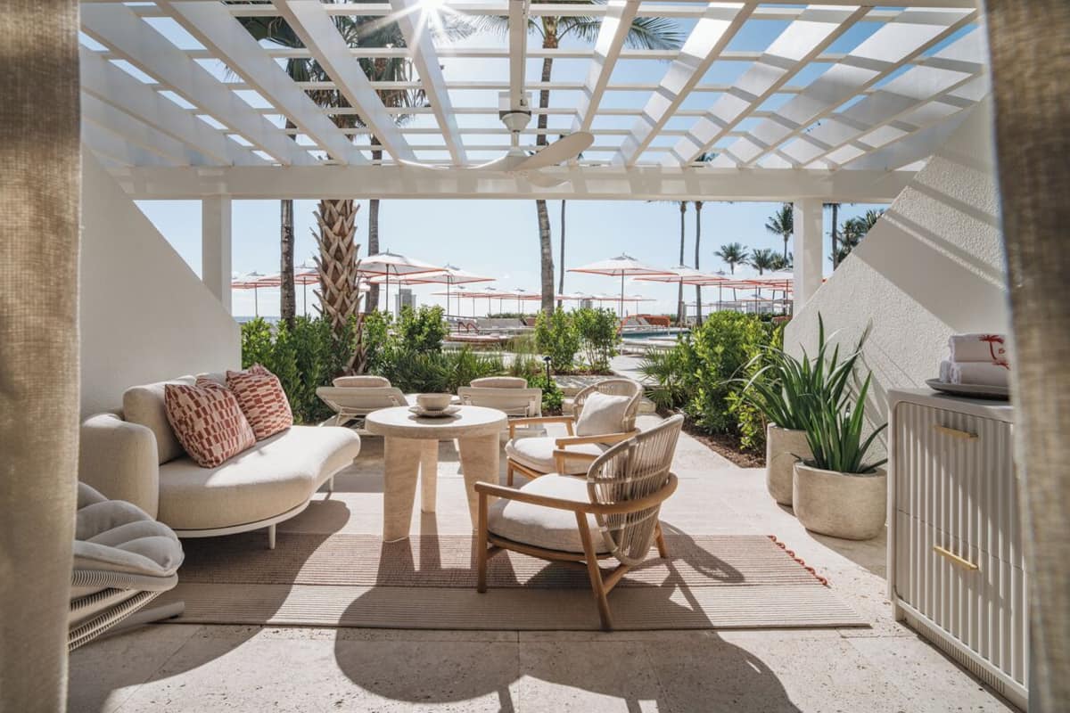 A sunlit outdoor seating area with a white pergola, lounge chairs, a small table, and potted plants.