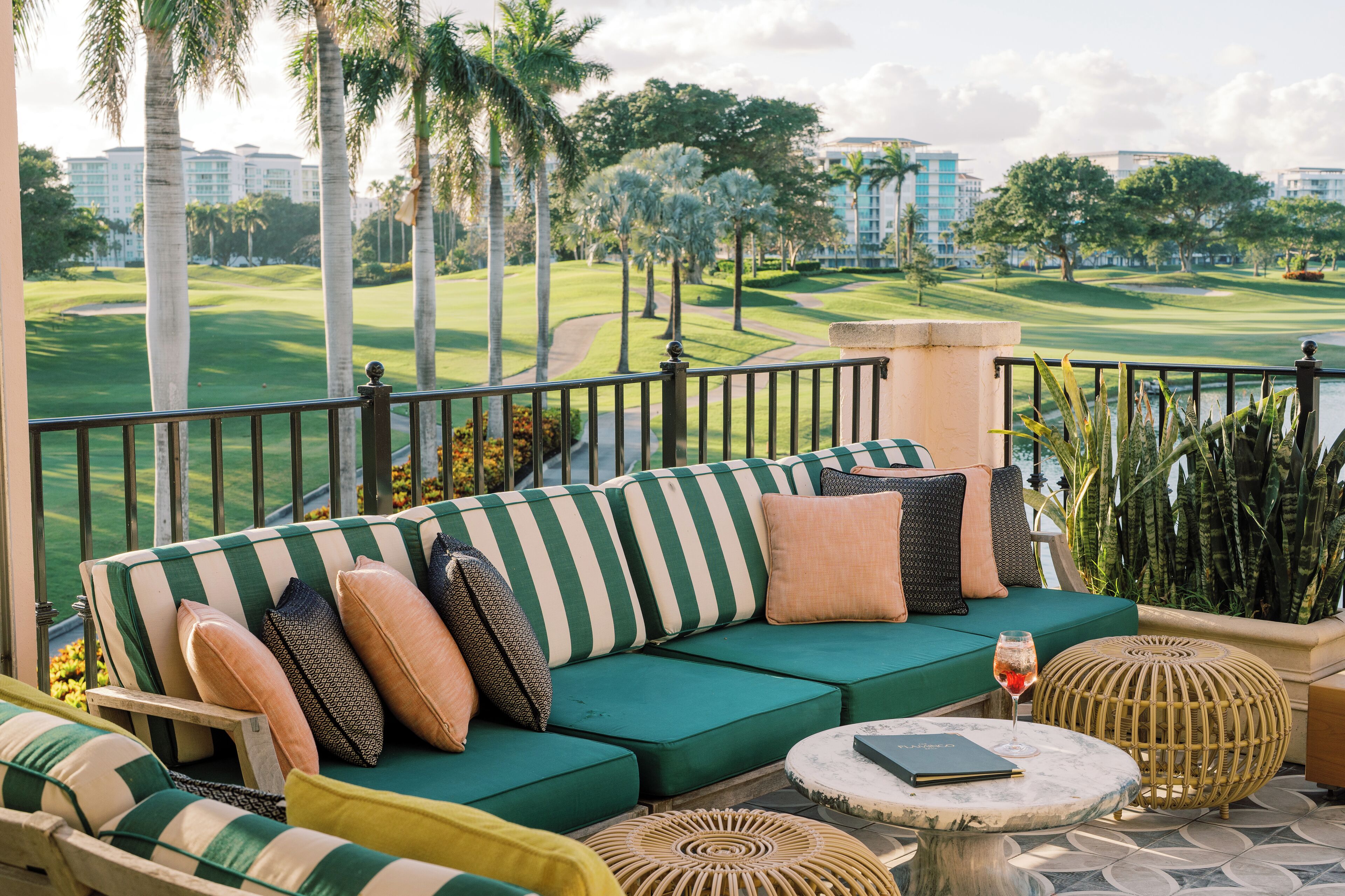 A green-striped sofa with cushions, a round table, and a wicker ottoman on a balcony with a view of a golf course and palm trees.