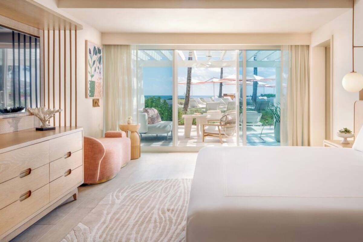 A modern hotel room with a large bed, a desk, a chair, and a balcony with outdoor seating.