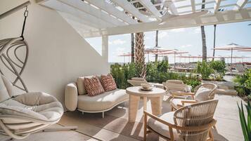 A sunlit outdoor lounge area with a white pergola, cushioned seating, and a view of the pool and umbrellas.