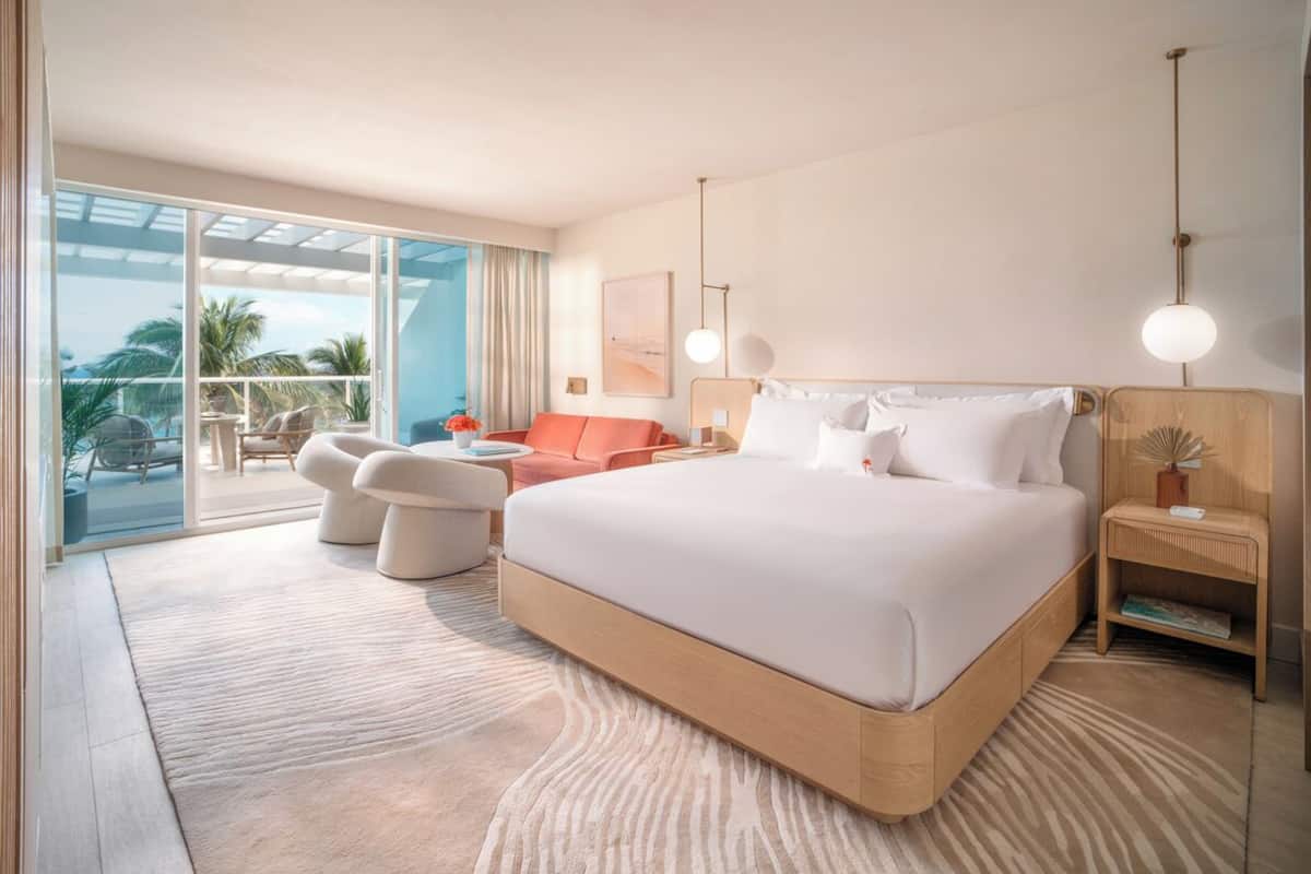 A modern hotel room with a large bed, a seating area with a sofa and chairs, a small table, and a balcony with a view of palm trees.