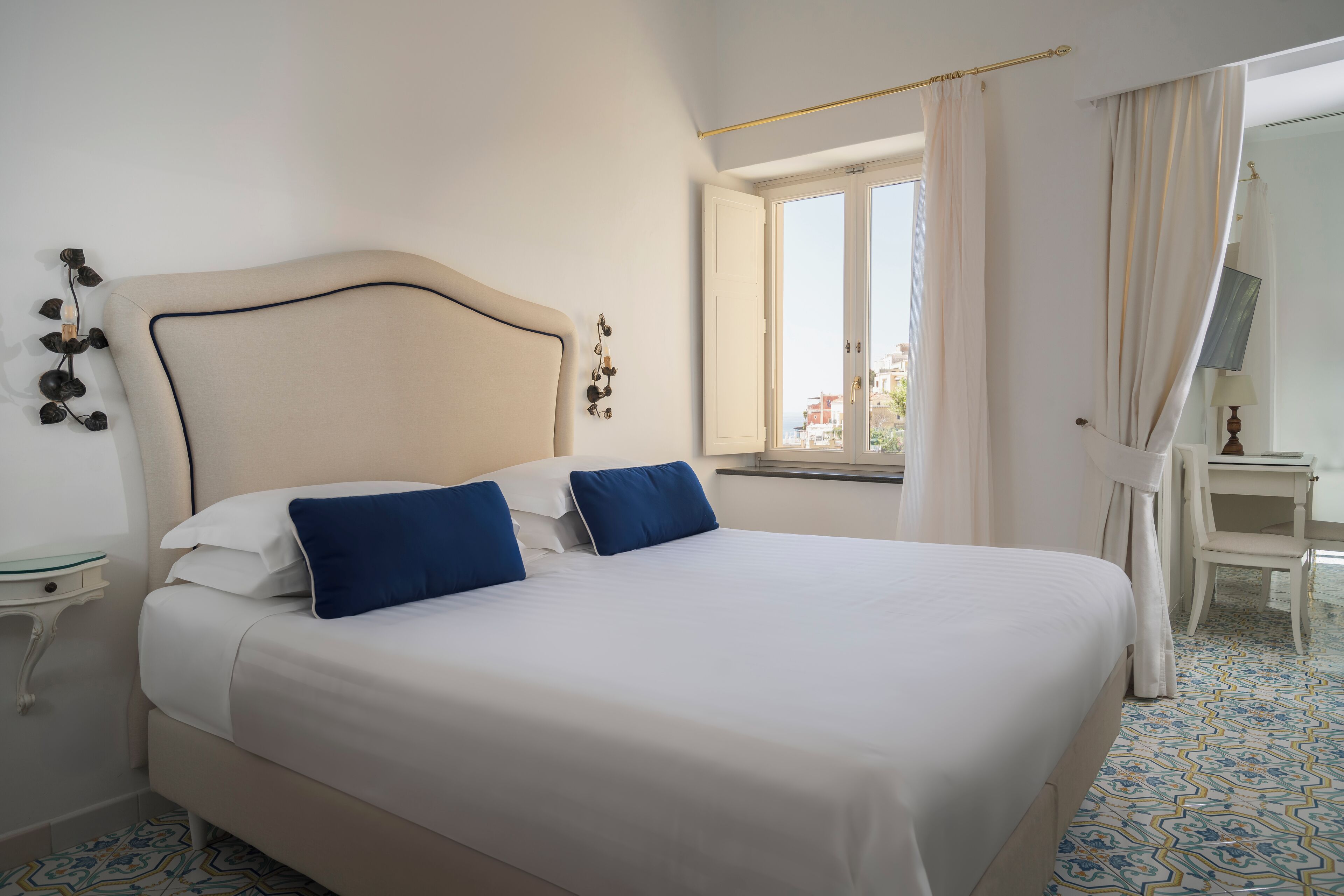 Une chambre d’hôtel avec un grand lit, deux oreillers, une table de chevet, une petite table, une fenêtre avec des rideaux et un sol à motifs.