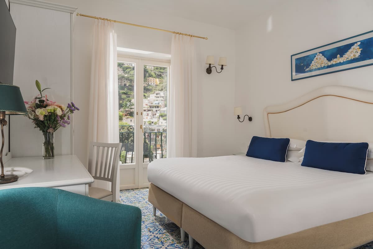 Une chambre d’hôtel avec un lit, un bureau, une chaise, un vase de fleurs, un tableau et une vue sur les bâtiments par la fenêtre.