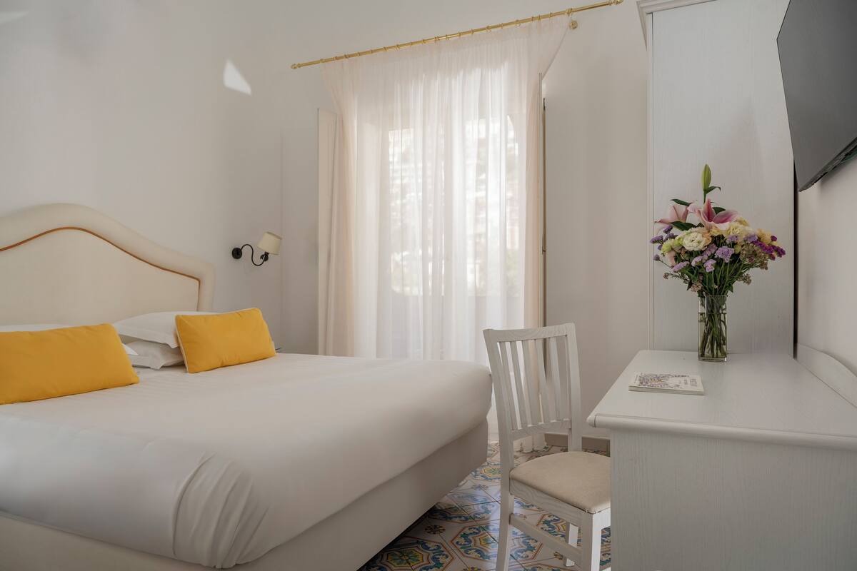 Une chambre d’hôtel avec un lit, un bureau, une chaise, un vase de fleurs et une fenêtre avec des rideaux.