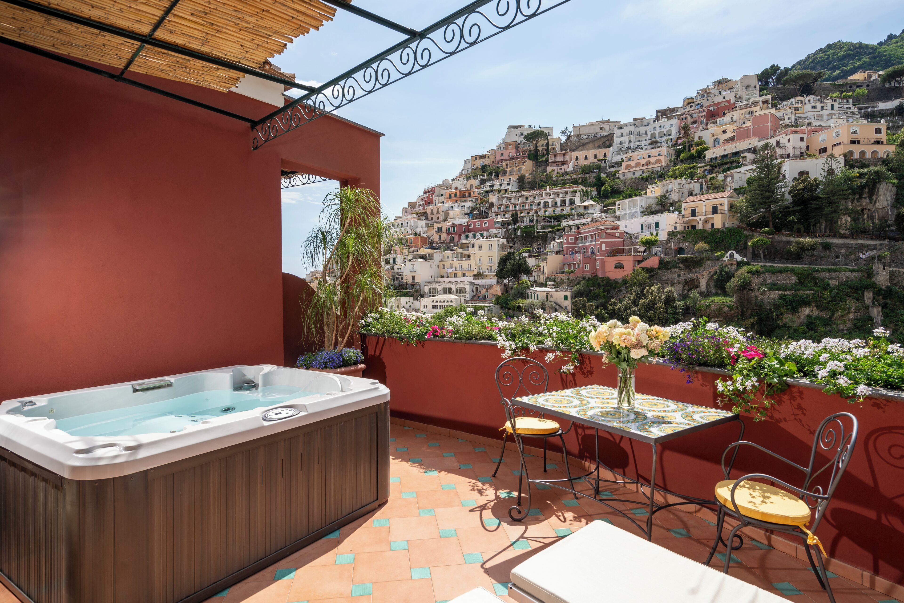 A rooftop terrace with a hot tub, a table with chairs, and a view of a hillside town.