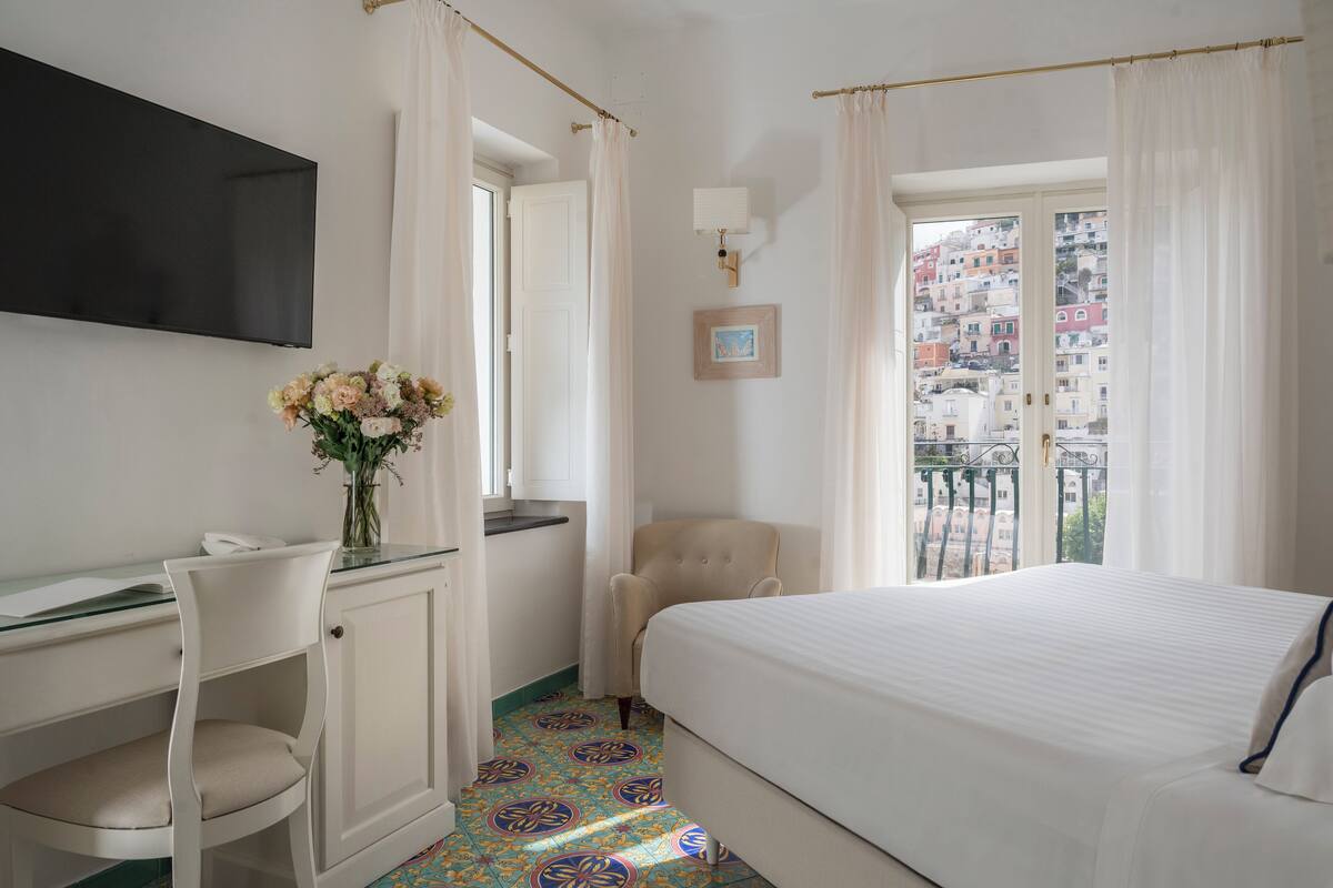 Une chambre d’hôtel comprenant un lit, un bureau avec une chaise, une télévision, une fenêtre avec des rideaux et une vue sur les bâtiments.