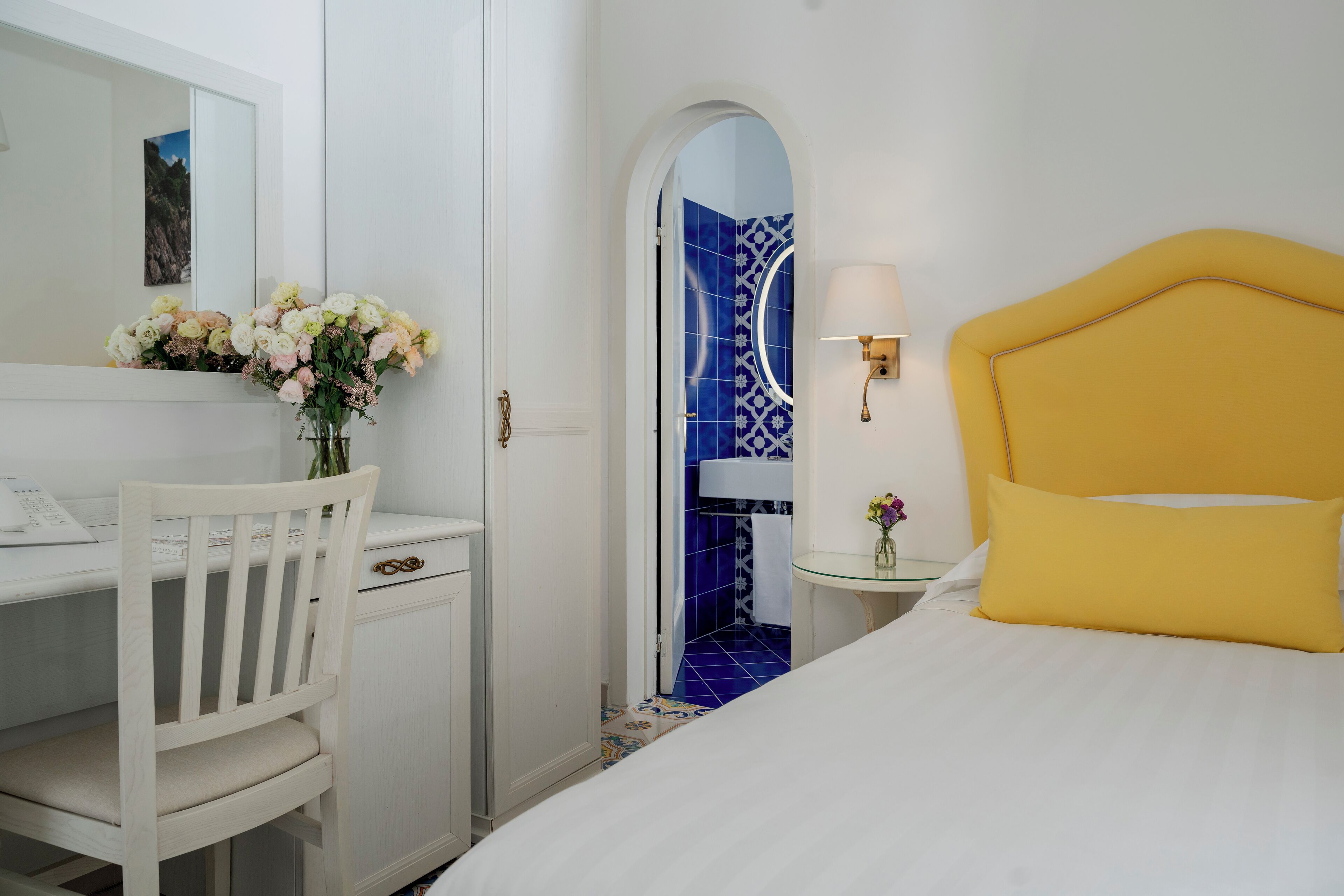A hotel room with a bed, a desk, and a chair. There is a mirror above the desk and a lamp on the bedside table.