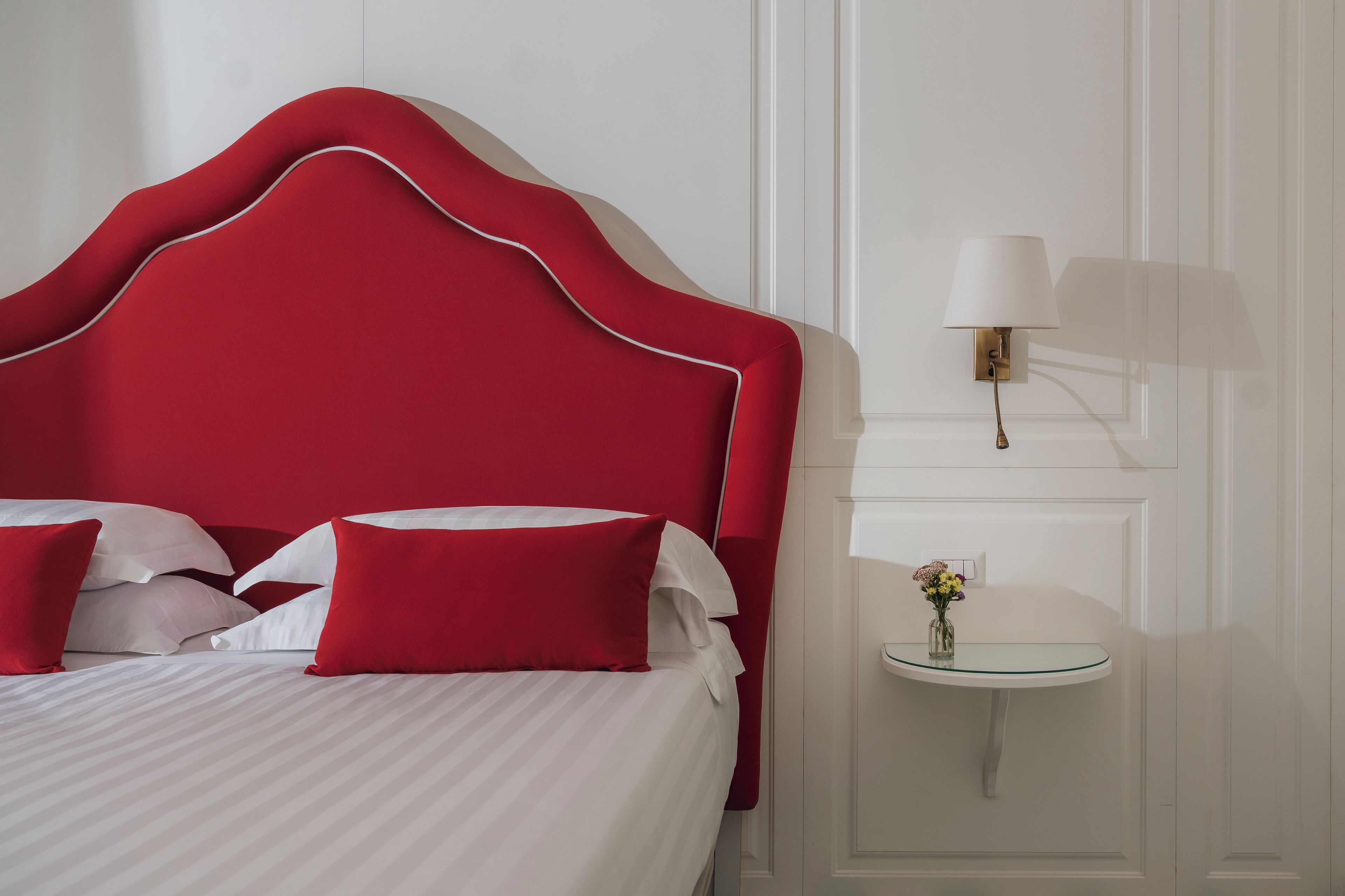 A neatly made bed with red and white bedding, a red upholstered headboard, and a small bedside table with a lamp and a vase of flowers.