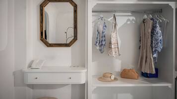 A white vanity with a mirror, a phone, and a small stool. Adjacent is a wardrobe with hanging clothes and a hat on a shelf.