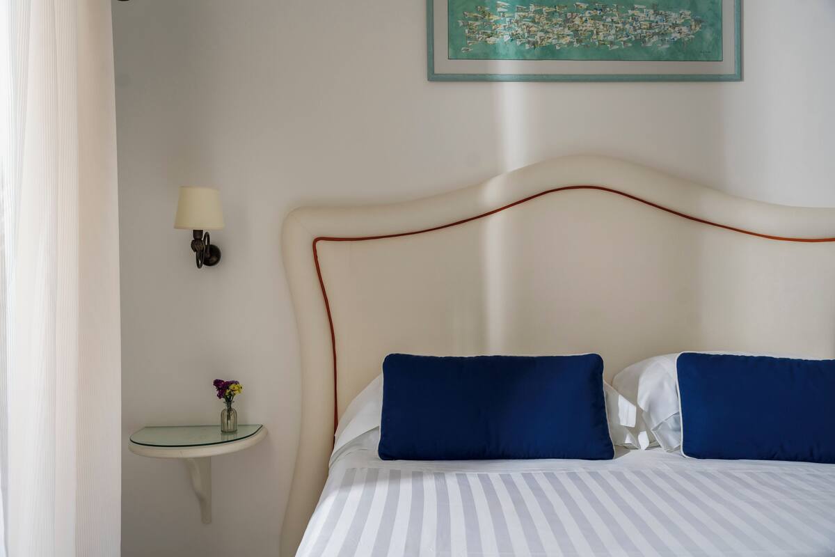 Un lit bien fait, orné de coussins bleus, une petite table de chevet avec un vase de fleurs et une lampe fixée au mur.