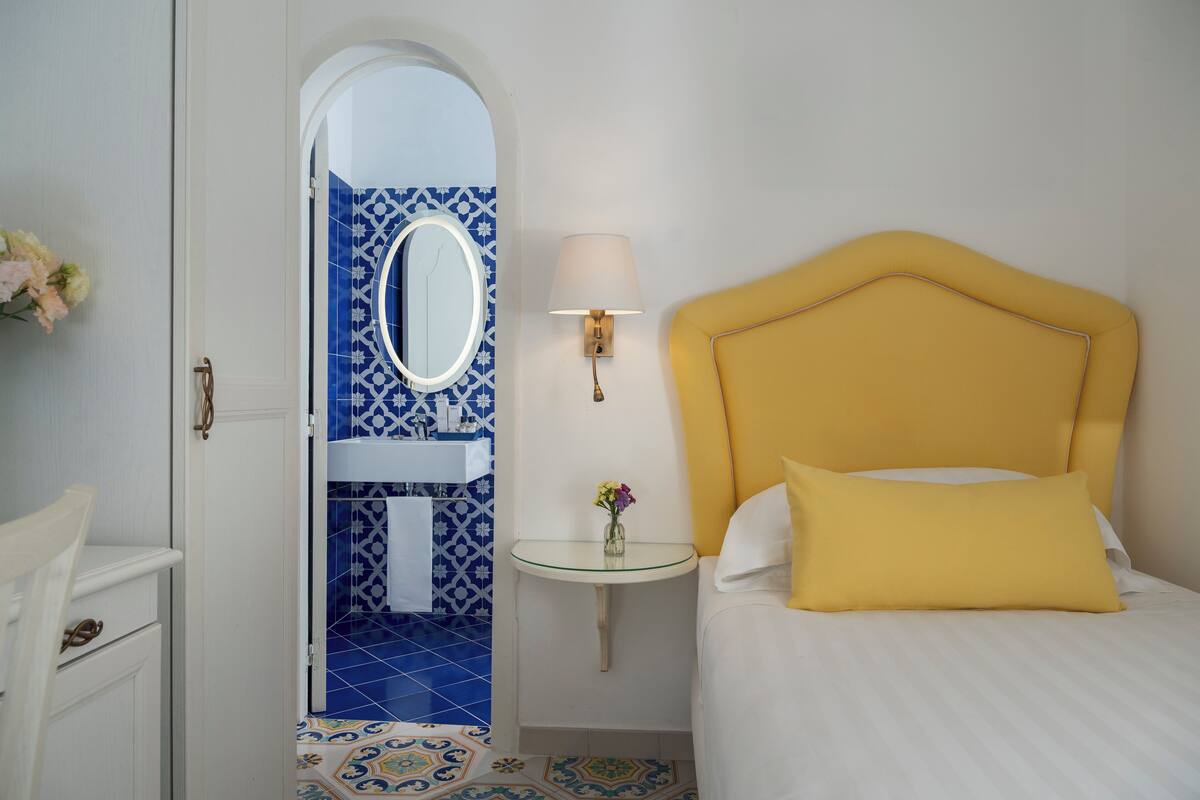 Une chambre à coucher avec une tête de lit jaune, une table de chevet avec une lampe, un miroir et une salle de bain carrelée de motifs bleus.