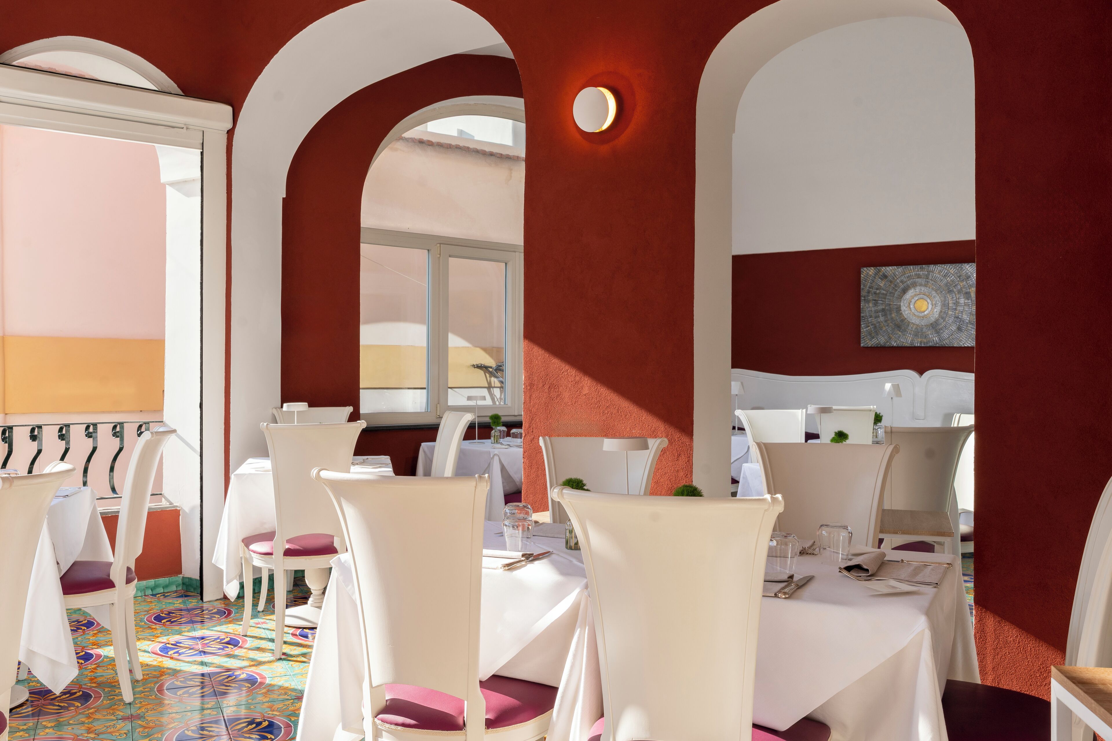 A dining area with white tables and chairs, a colorful floor, and a red wall with a circular light fixture.