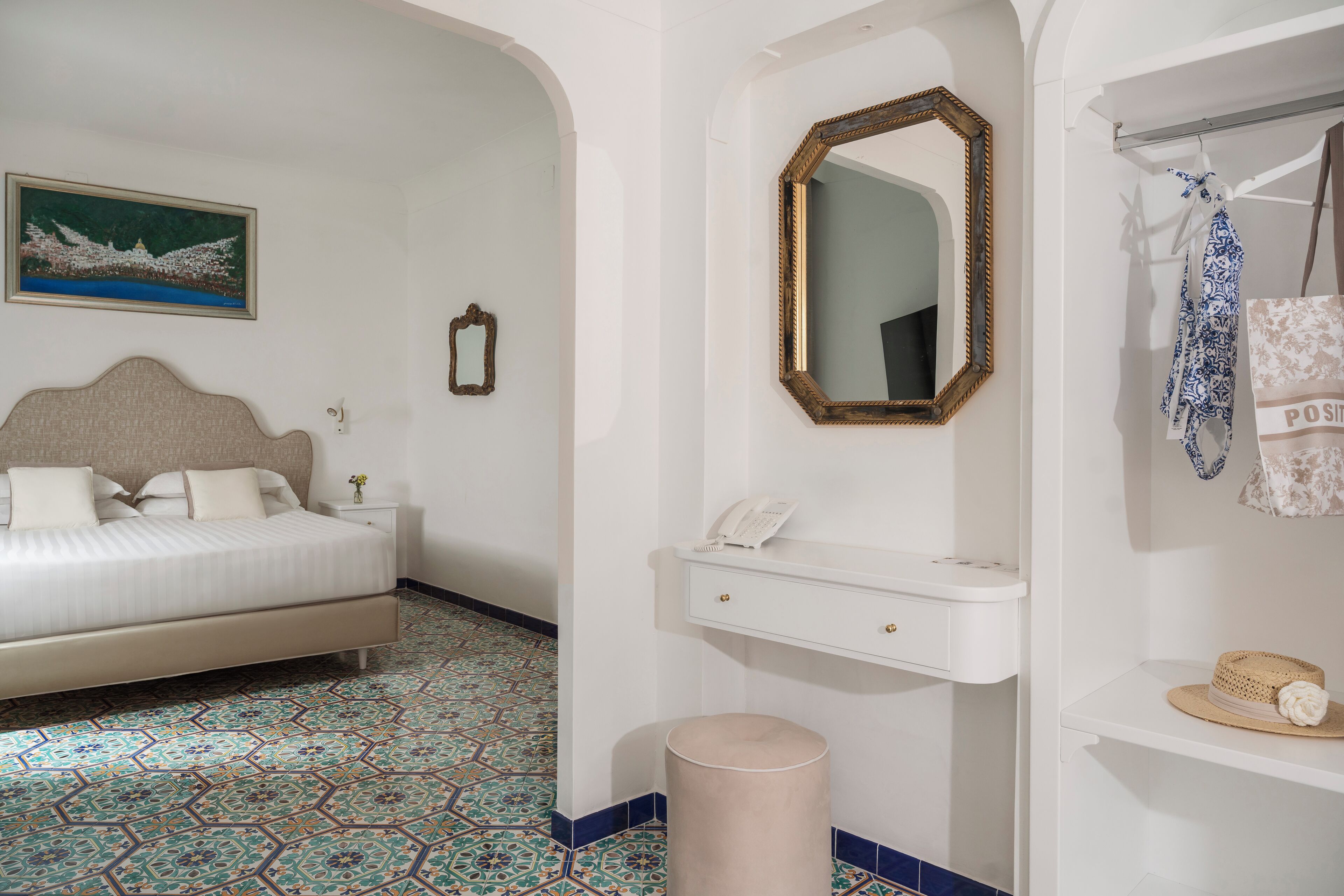 A bedroom with a bed, a bedside table with a lamp, a mirror, a vanity with a phone, and a wall-mounted shelf with a hat.